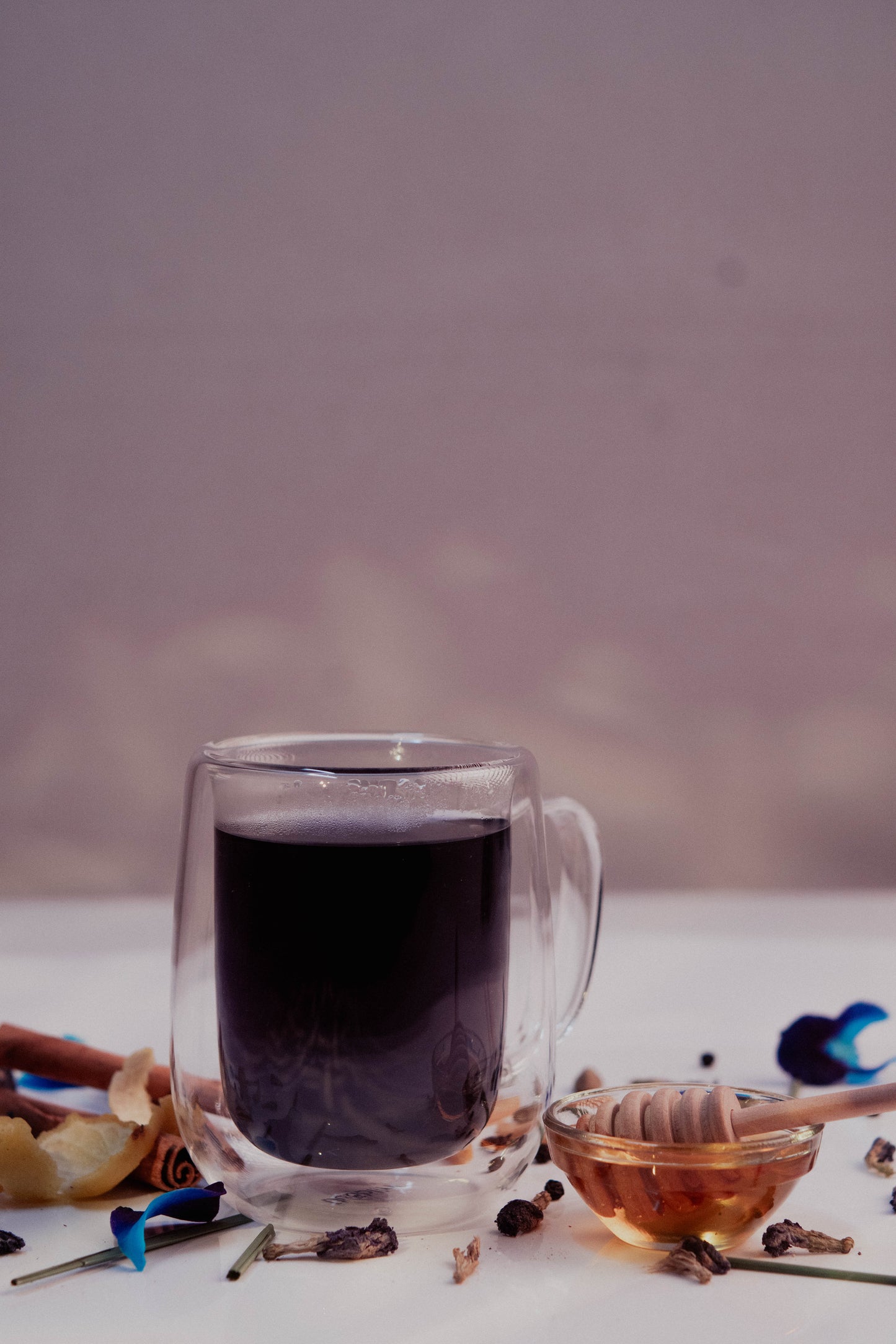 Clear mug with dark liquid on a white surface with honey and spices.