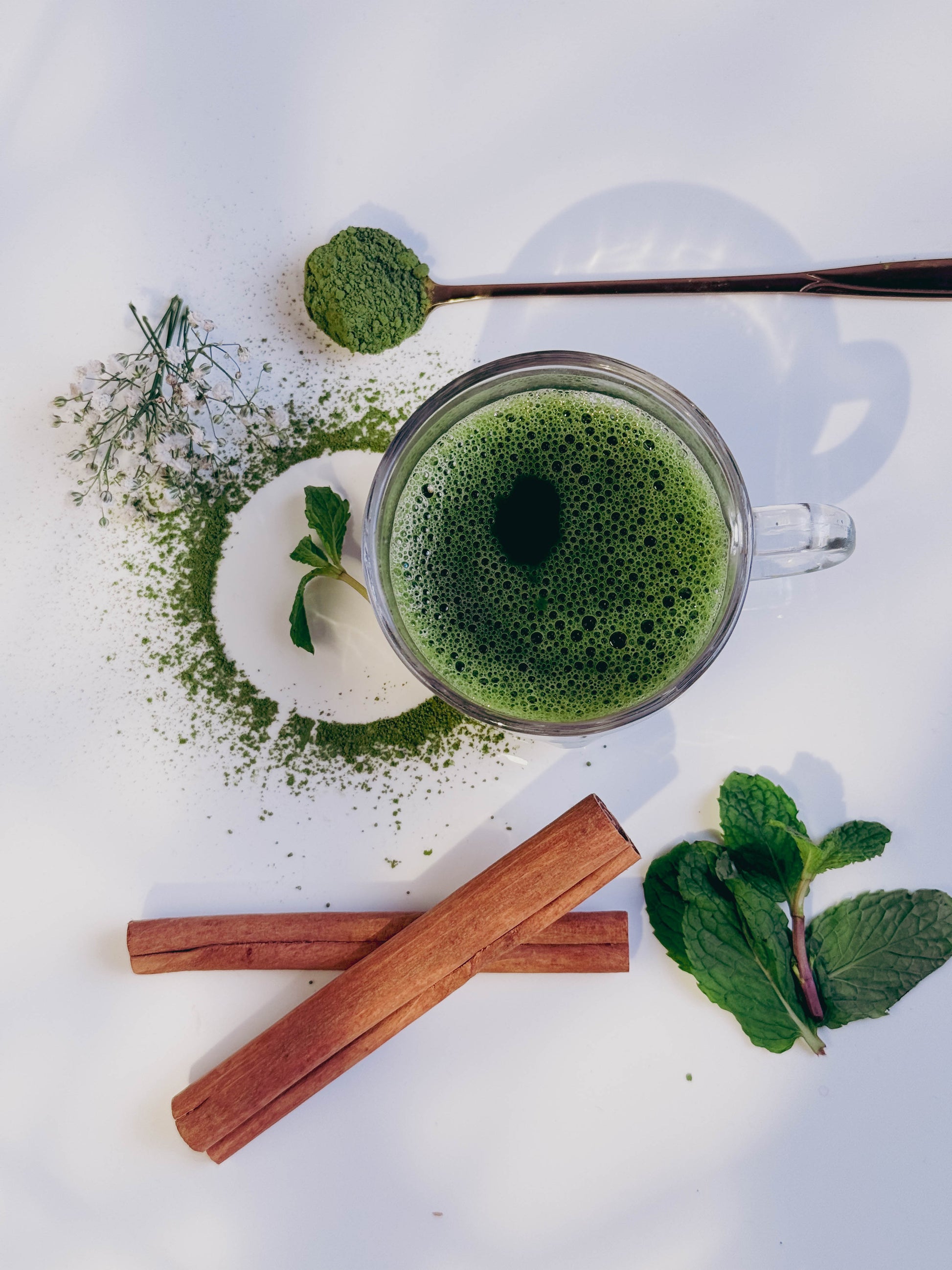 Clear mug filled with green matcha tea, surrounded by matcha powder, mint leaves, and cinnamon sticks on a white background.